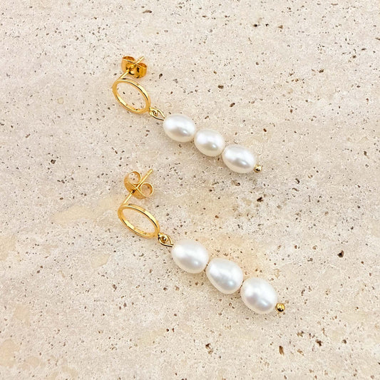 A pair of Aking Sinta gold triple pearl drop earrings. Each earring features three white freshwater pearls suspended from a polished gold circle stud, laid out on a natural stone surface