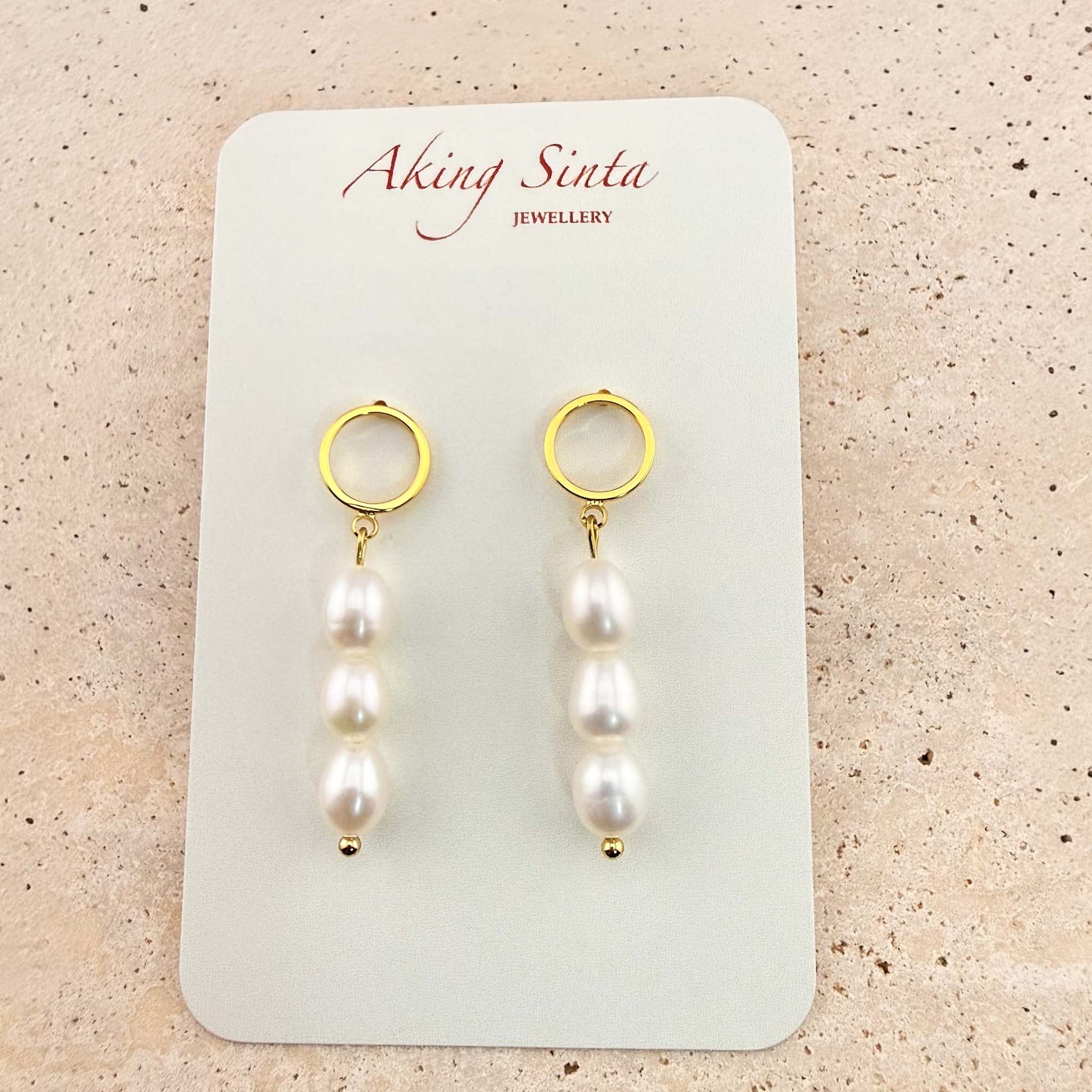 A pair of Aking Sinta triple pearl drop earrings on a white branded display card. Each earring features three white oval pearls hanging from a gold circle stud, set against a light stone background