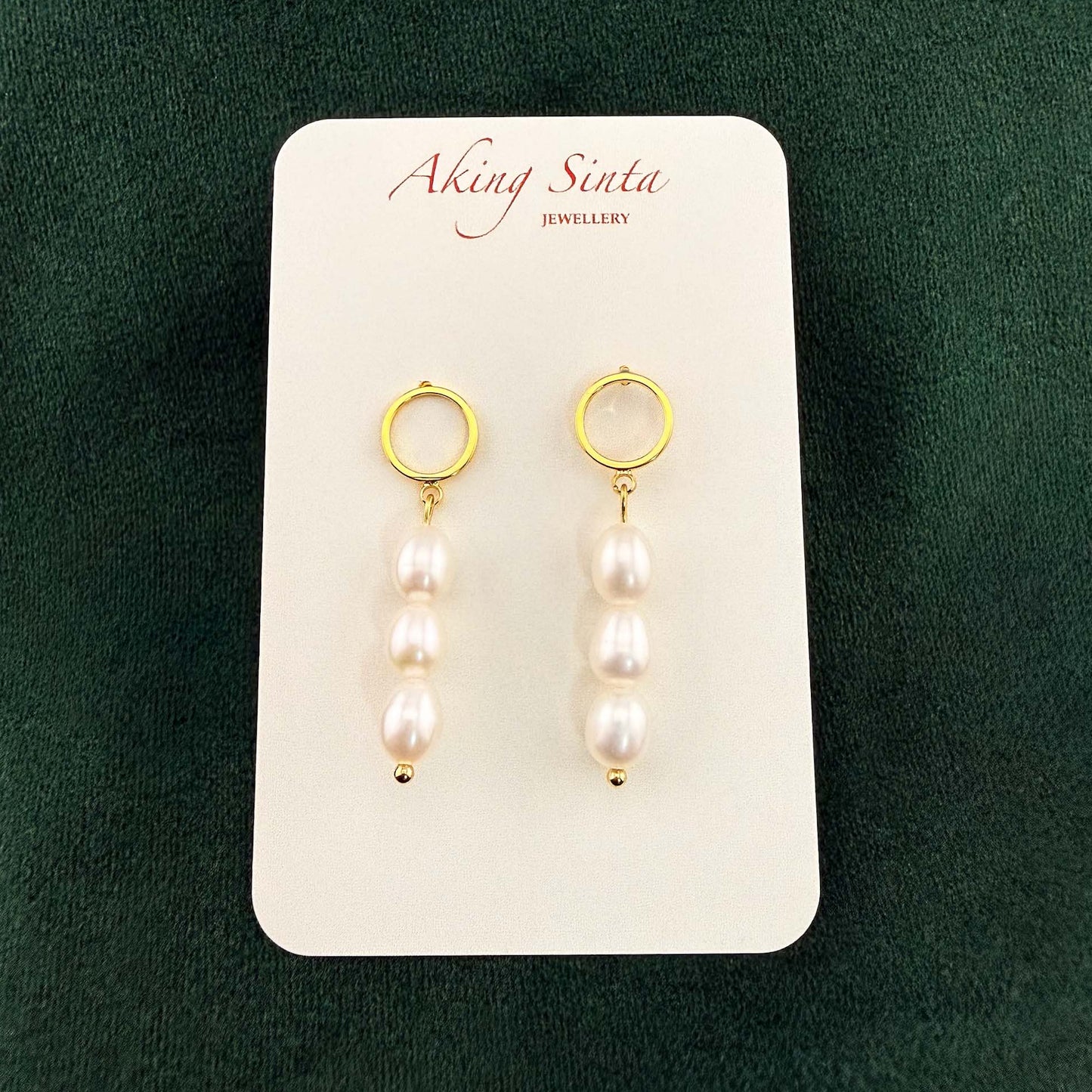 A pair of Aking Sinta triple pearl drop earrings. Each earring features three white oval pearls dangling from a minimalist gold circle stud, displayed on a branded white card against a dark green velvet background