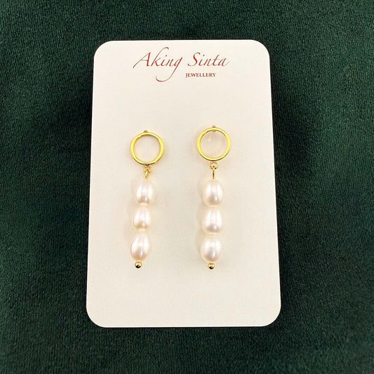 A pair of Aking Sinta triple pearl drop earrings. Each earring features three white oval pearls dangling from a minimalist gold circle stud, displayed on a branded white card against a dark green velvet background