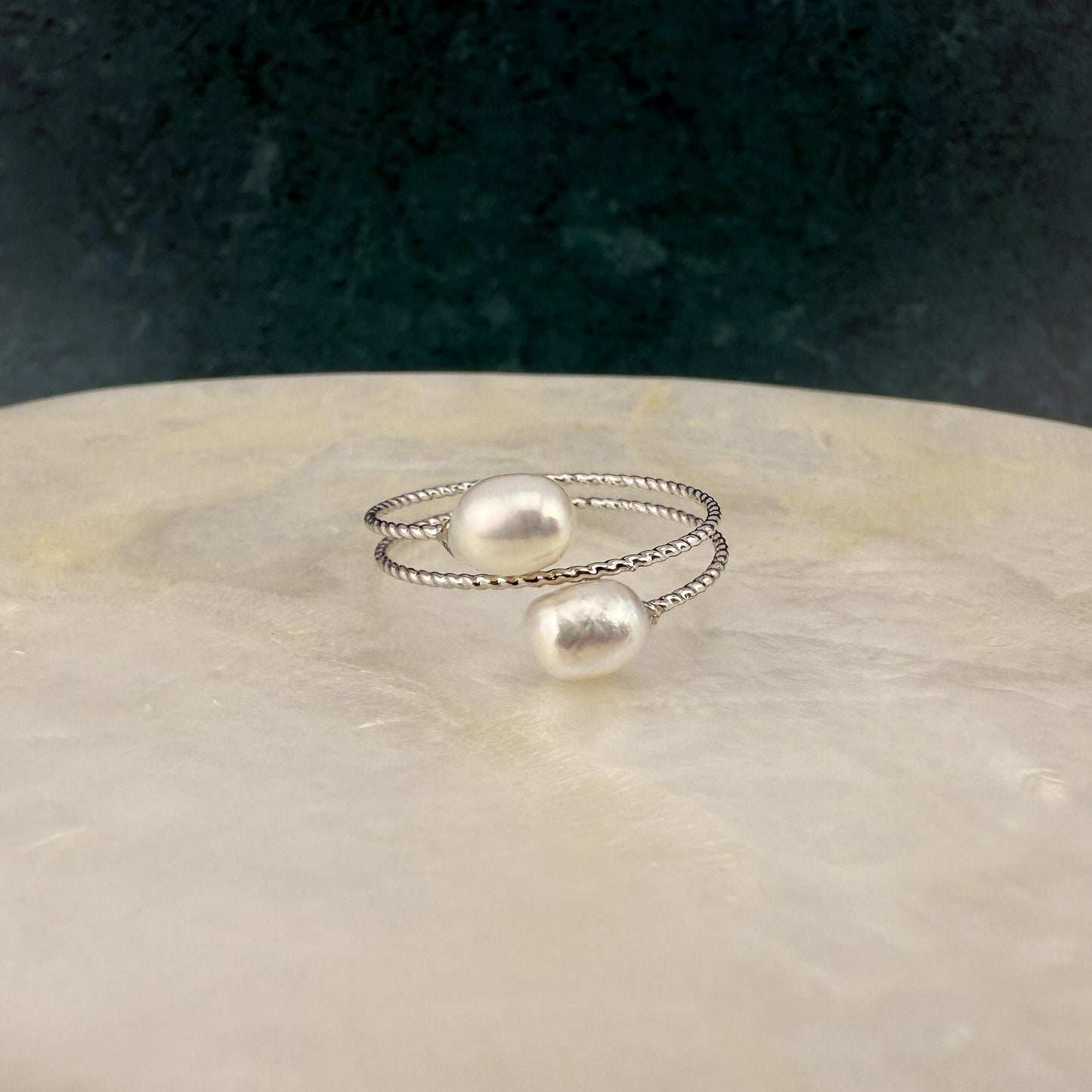 Close-up of an Aking Sinta white double pearl adjustable ring on a white marble slab; featuring two classic creamy pearls on a dainty gold band against a dark green background.
