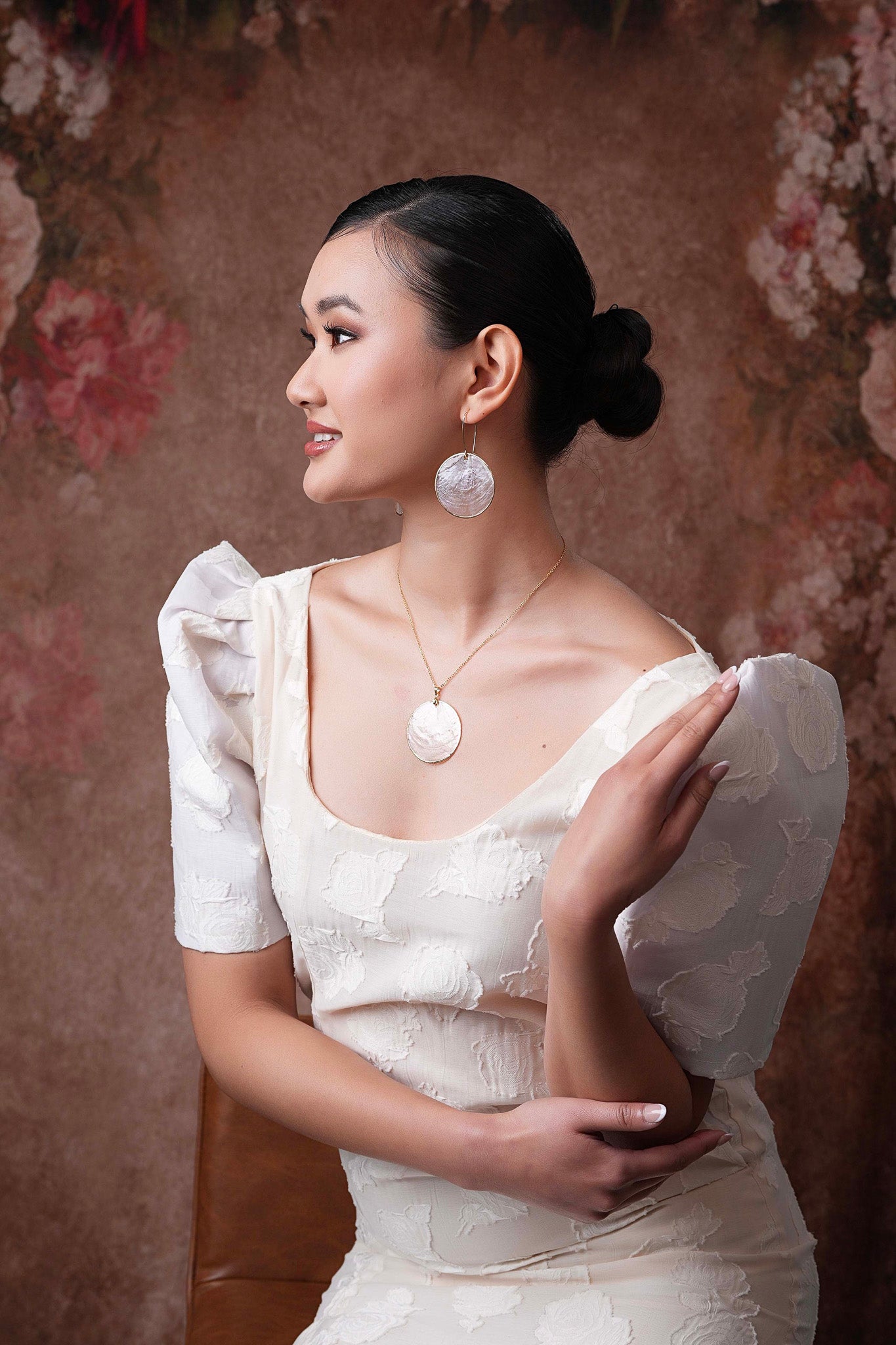 Side view of a model wearing a white top and Zoey’s Capiz Shell Wedding Earrings; the large, translucent white Capiz disk hangs from a gold hook, resting just above the shoulder and glowing against the model's skin and white clothing.