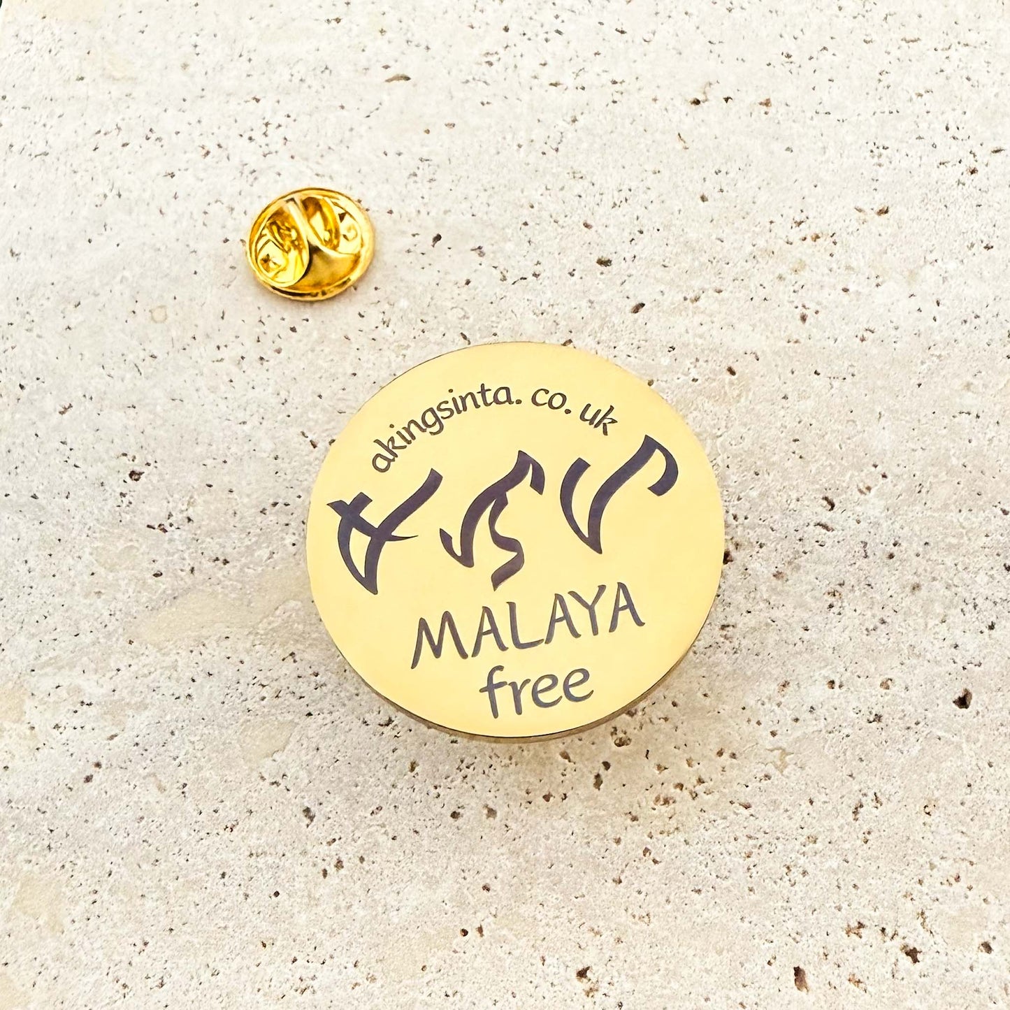 An Aking Sinta product photograph showcasing a circular gold lapel pin with its butterfly clutch back on a textured light stone surface. The polished pin is custom-engraved with the Aking Sinta brand website akingsinta.co.uk at the top, traditional Baybayin characters in the center, and the word "MALAYA" with its translation "free" below.