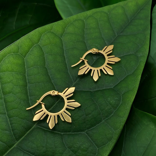A pair of Araw gold sun hoop earrings with 5-ray detailing, laid on a textured green tropical leaf for a nature-inspired jewelry display.