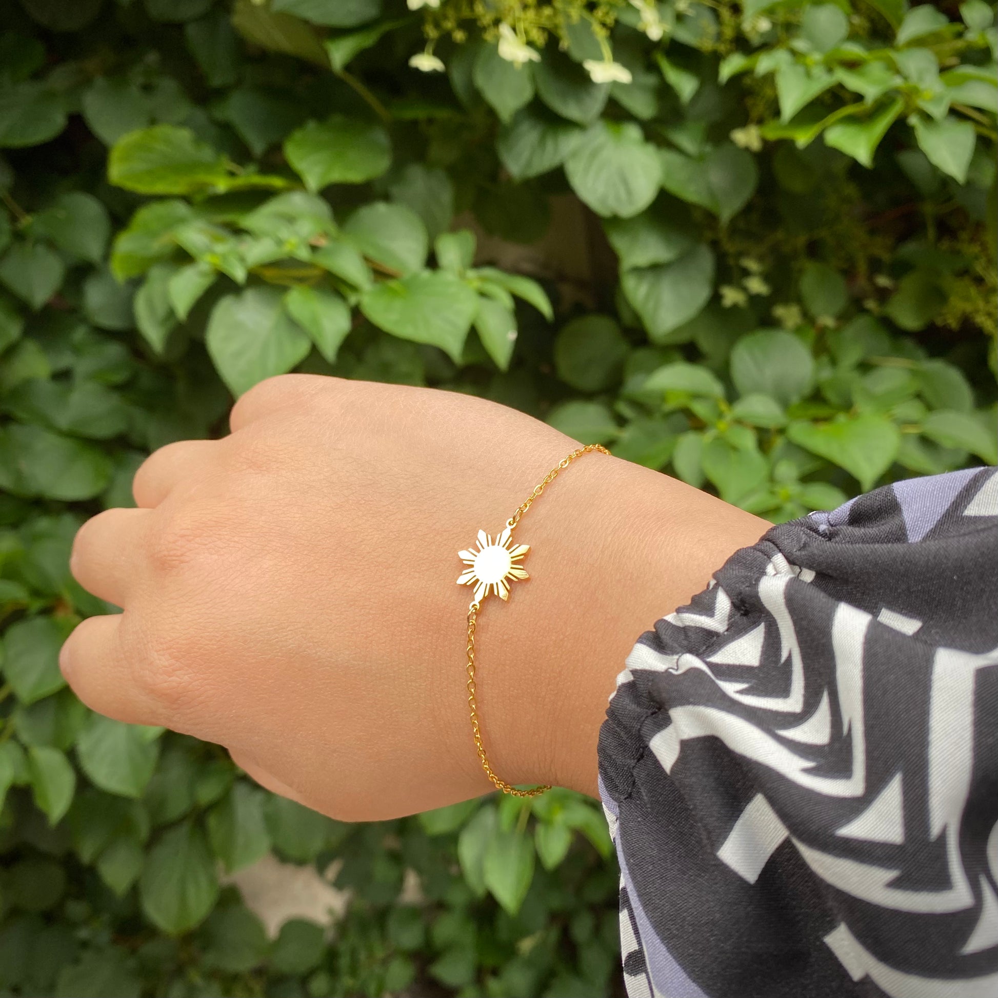 Close up of a hand wearing the Araw minimalist gold and silver sun jewelry with a soft bokeh background of green flowering plants and lush garden foliage