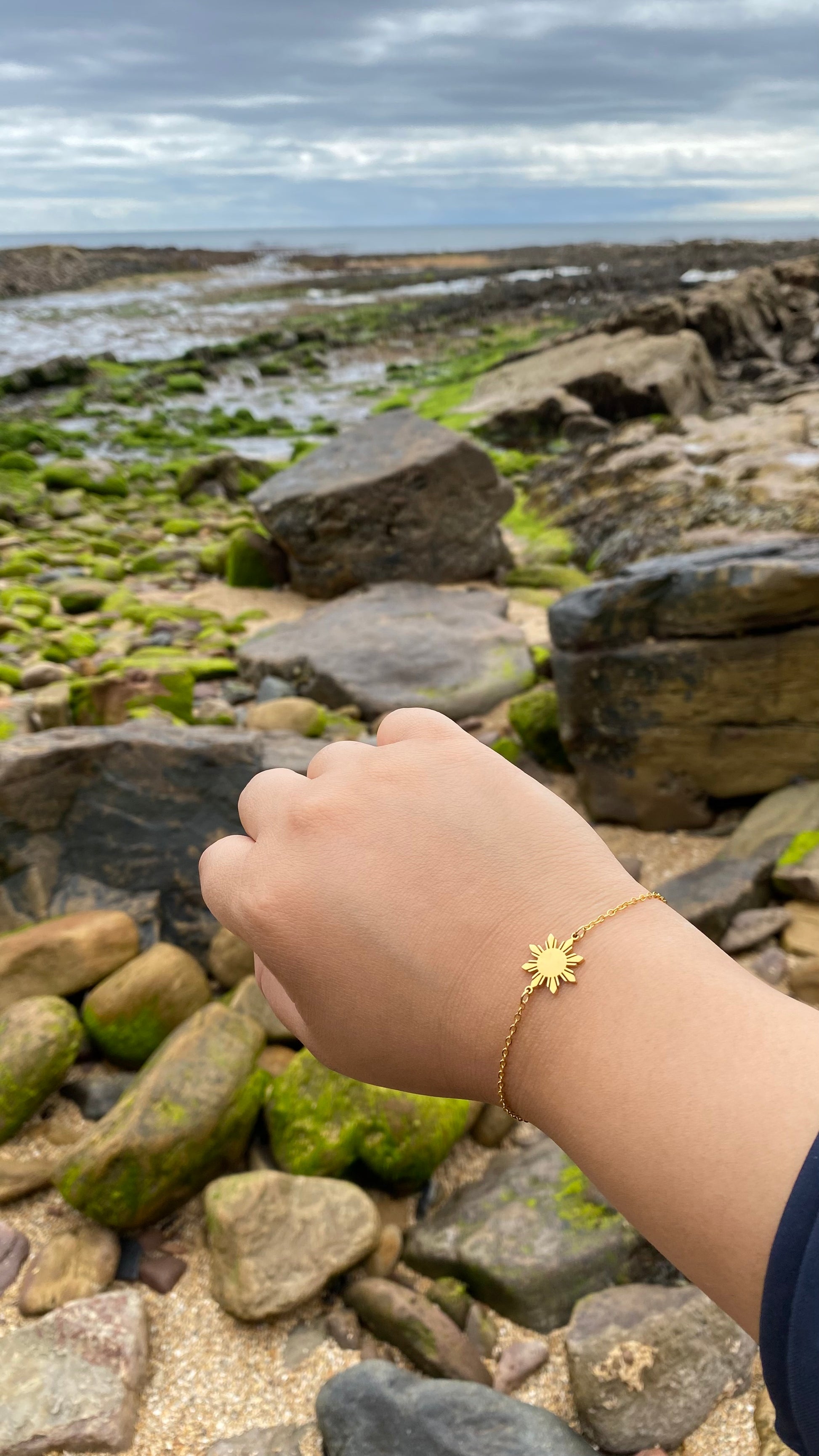 Close up of a hand wearing the Araw minimalist sun jewelry with a blurred beach and ocean background, capturing a summer vacation lifestyle aesthetic