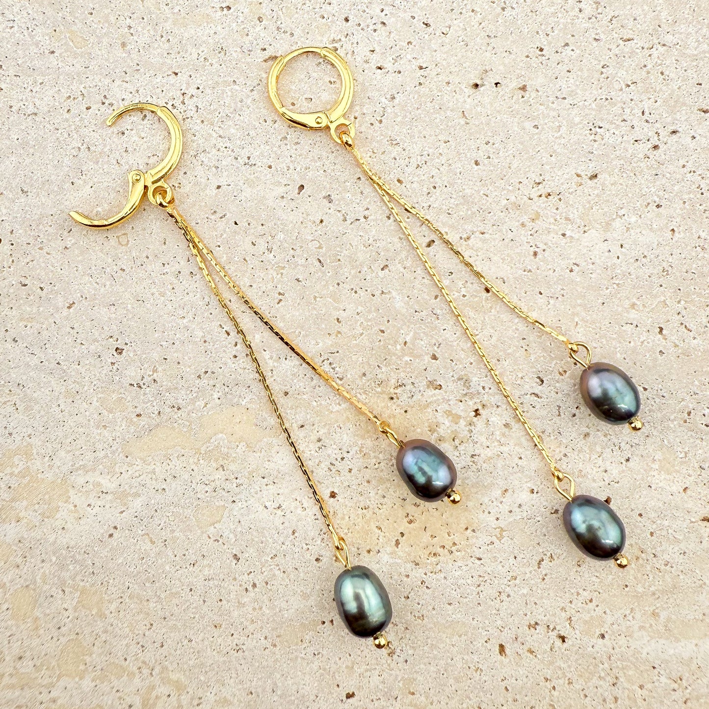 Macro view of a gold Pearl Double Drop earring on white marble, featuring two stacked black pearls with one hinge clasp securely open.