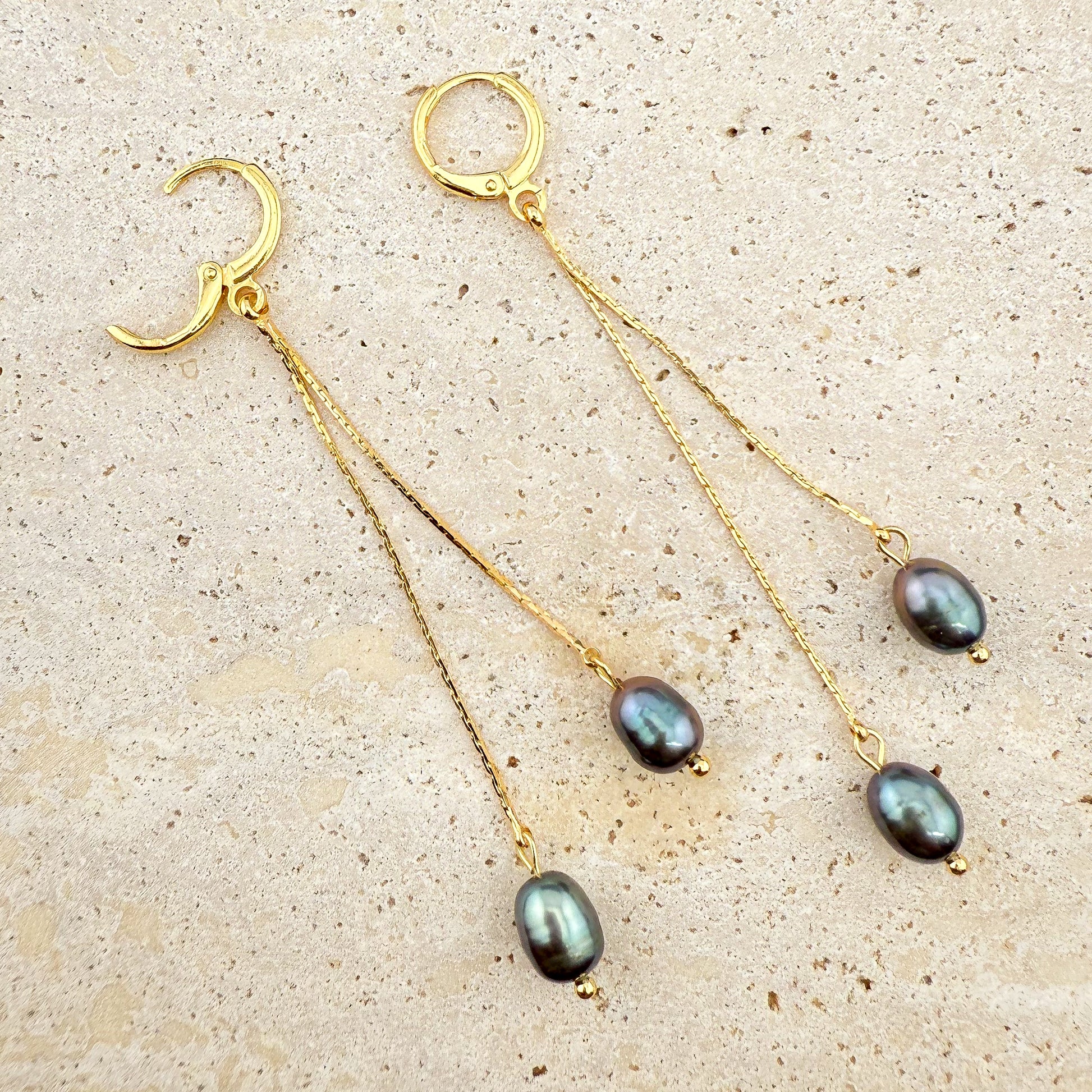 Macro view of a gold Pearl Double Drop earring on white marble, featuring two stacked black pearls with one hinge clasp securely open.
