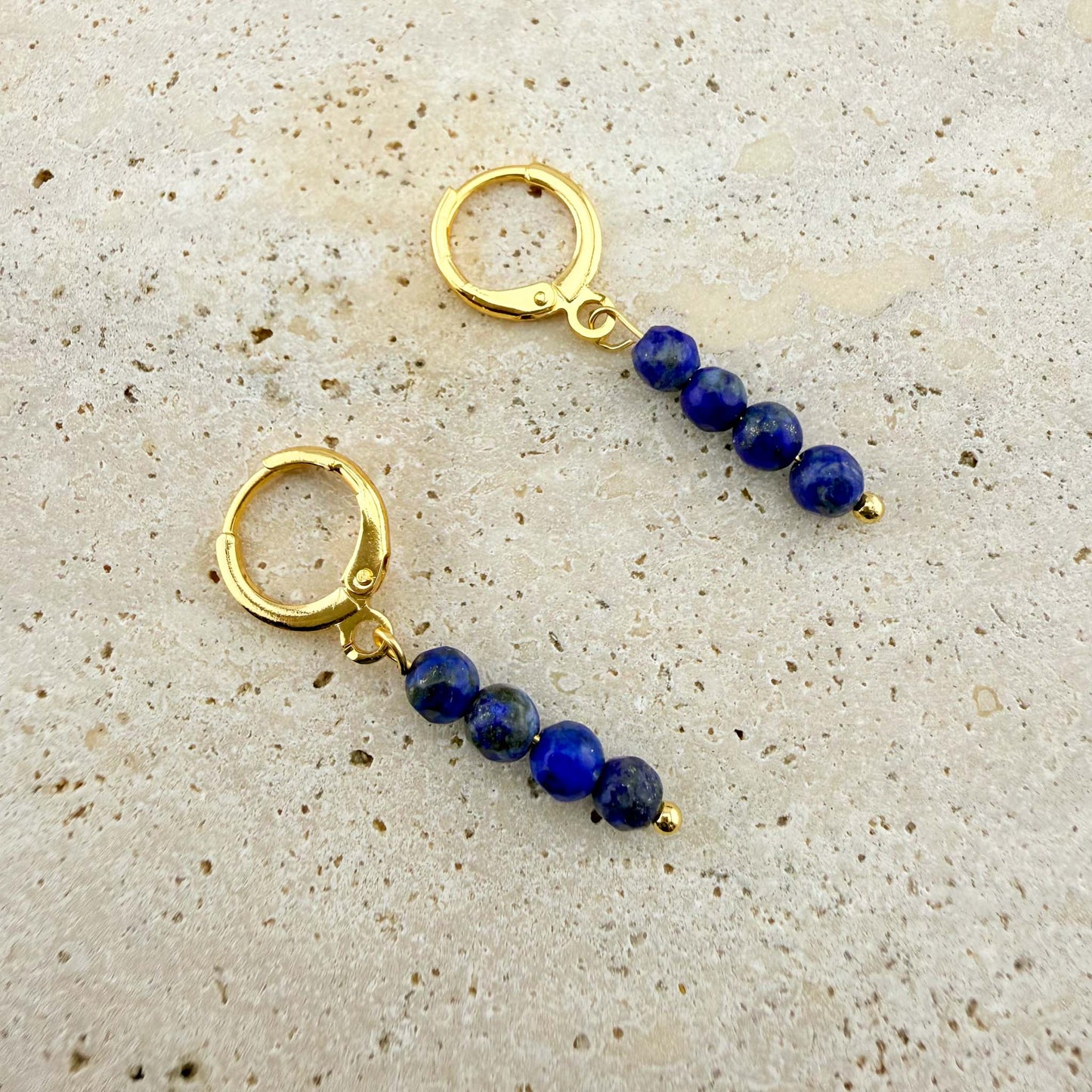 Macro view of gold Aking Sinta Lapis Lazuli Huggies on a white marble texture, featuring three stacked natural deep blue beads on each high-polish click-to-close hoop.