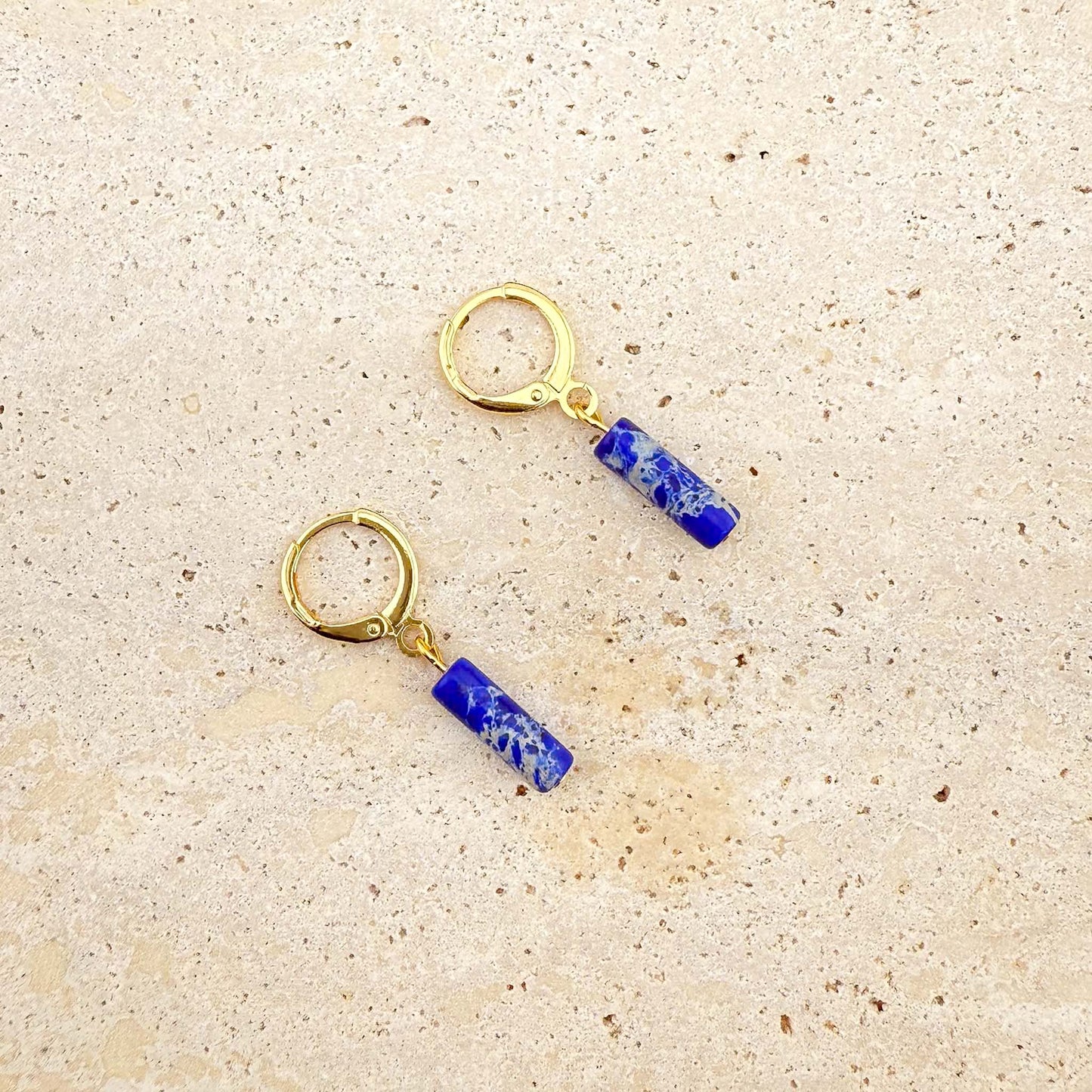 Close-up of gold and lapis lazuli cylinder drop earrings on beige stone; the upper earring's lever back clasp is open to show the mechanism, the lower is closed.