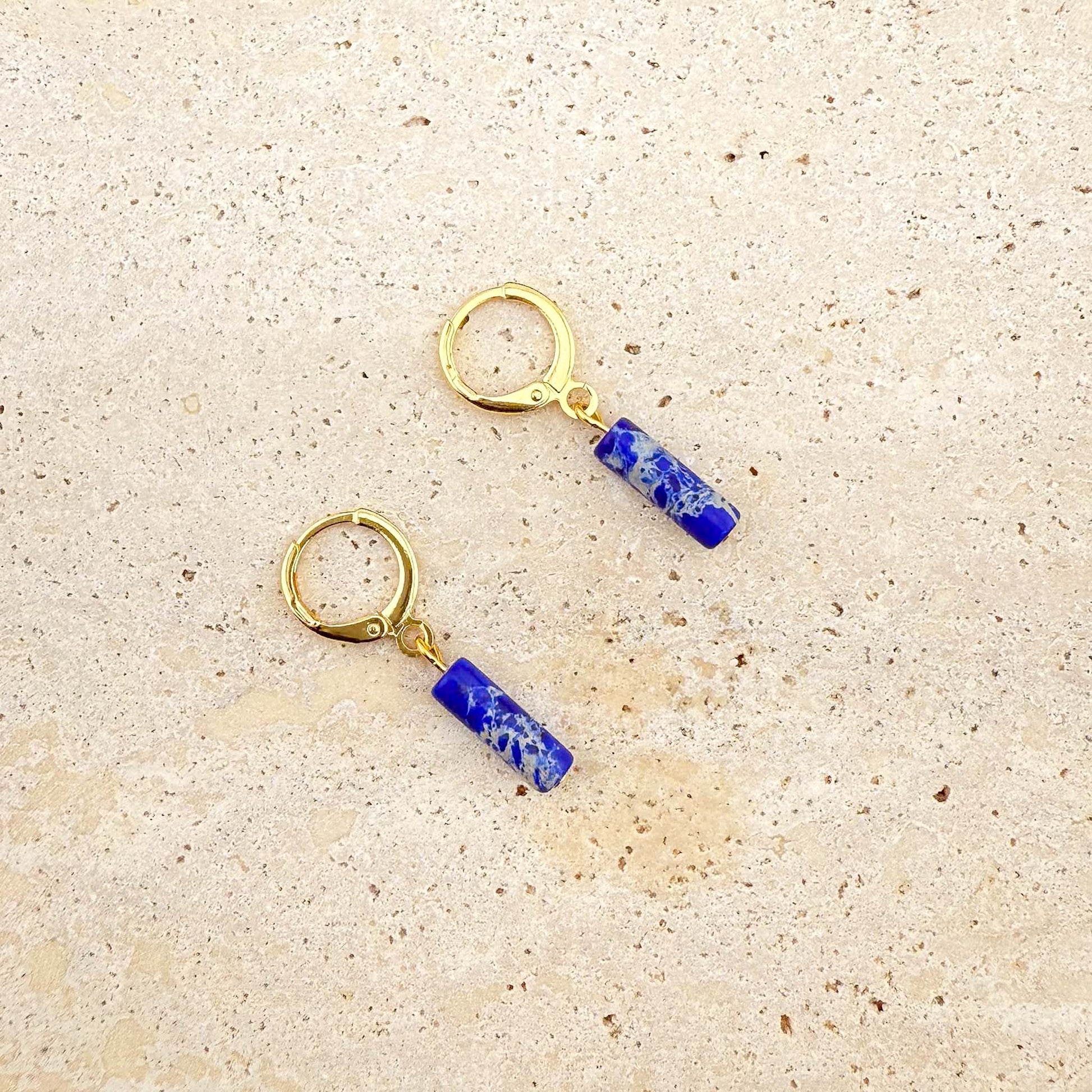Close-up of gold and lapis lazuli cylinder drop earrings on beige stone; the upper earring's lever back clasp is open to show the mechanism, the lower is closed.