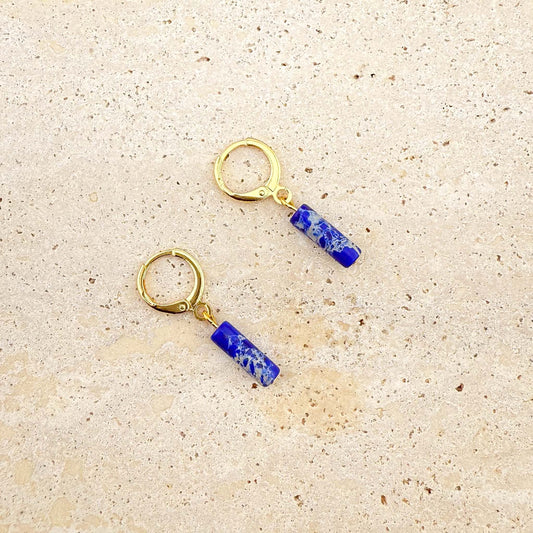 Close-up of gold and lapis lazuli cylinder drop earrings on beige stone; the upper earring's lever back clasp is open to show the mechanism, the lower is closed.