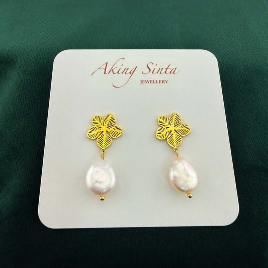 Dainty gold Sampaguita jasmine flower studs with small white coin pearl charms, pinned to a kraft cardboard jewelry holder on a dark forest green background.
