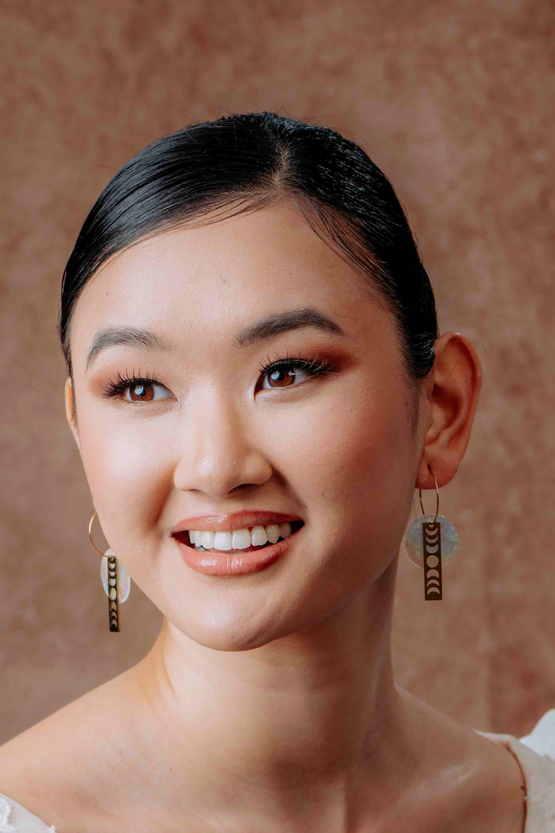 A close-up side view of a model wearing Aking Sinta gold bar earrings; the long, slim gold bar features vertical moon phase cutouts and ends with a dainty, round white Capiz shell charm that dangles just above the shoulder.
