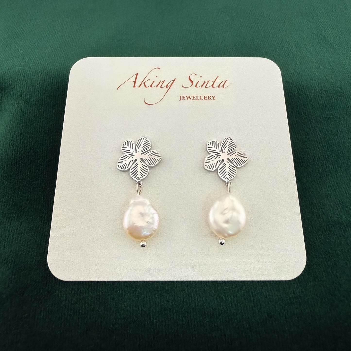 Dainty silver Sampaguita jasmine flower studs with small white coin pearl charms, pinned to a kraft cardboard jewelry holder on a dark forest green background.