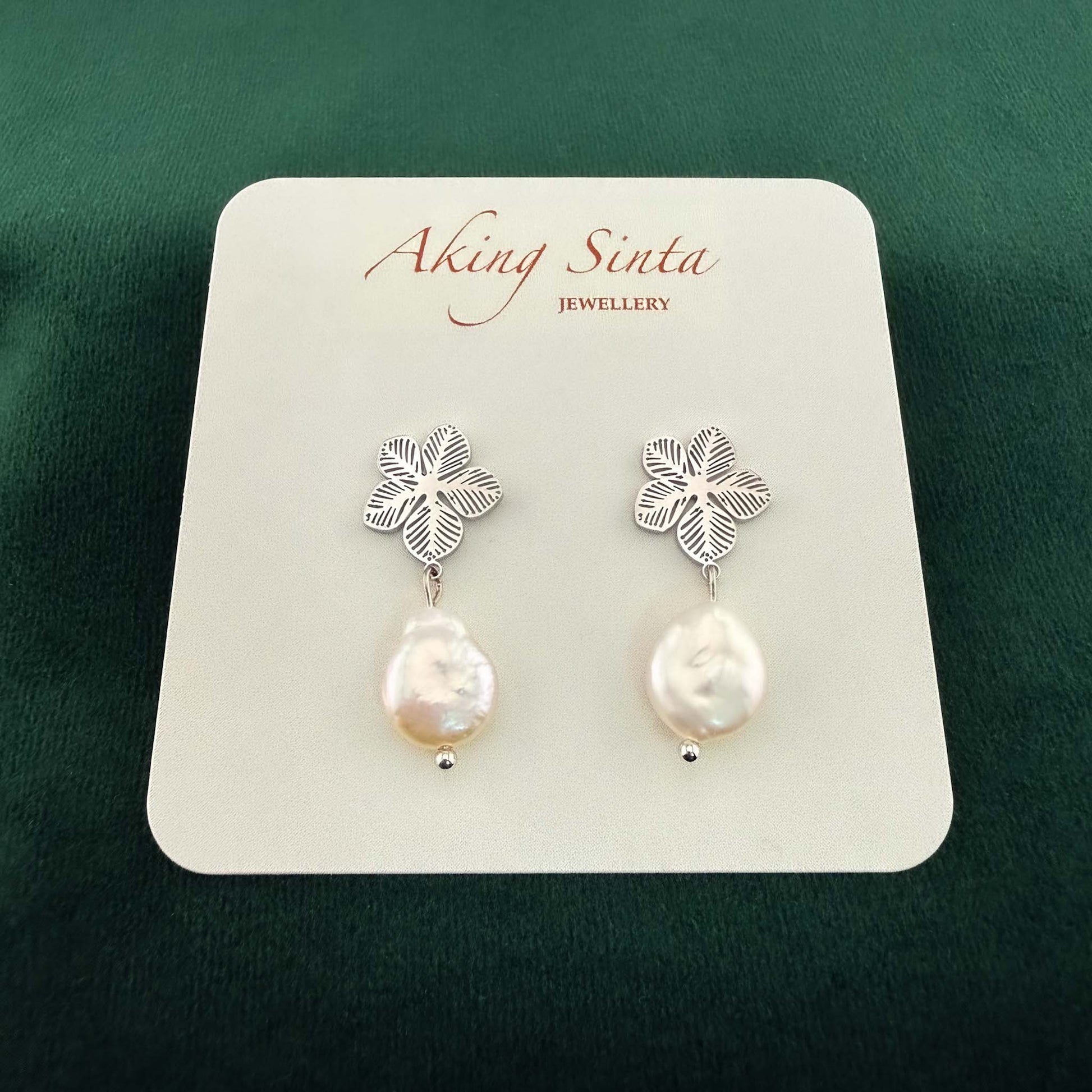 Dainty silver Sampaguita jasmine flower studs with small white coin pearl charms, pinned to a kraft cardboard jewelry holder on a dark forest green background.