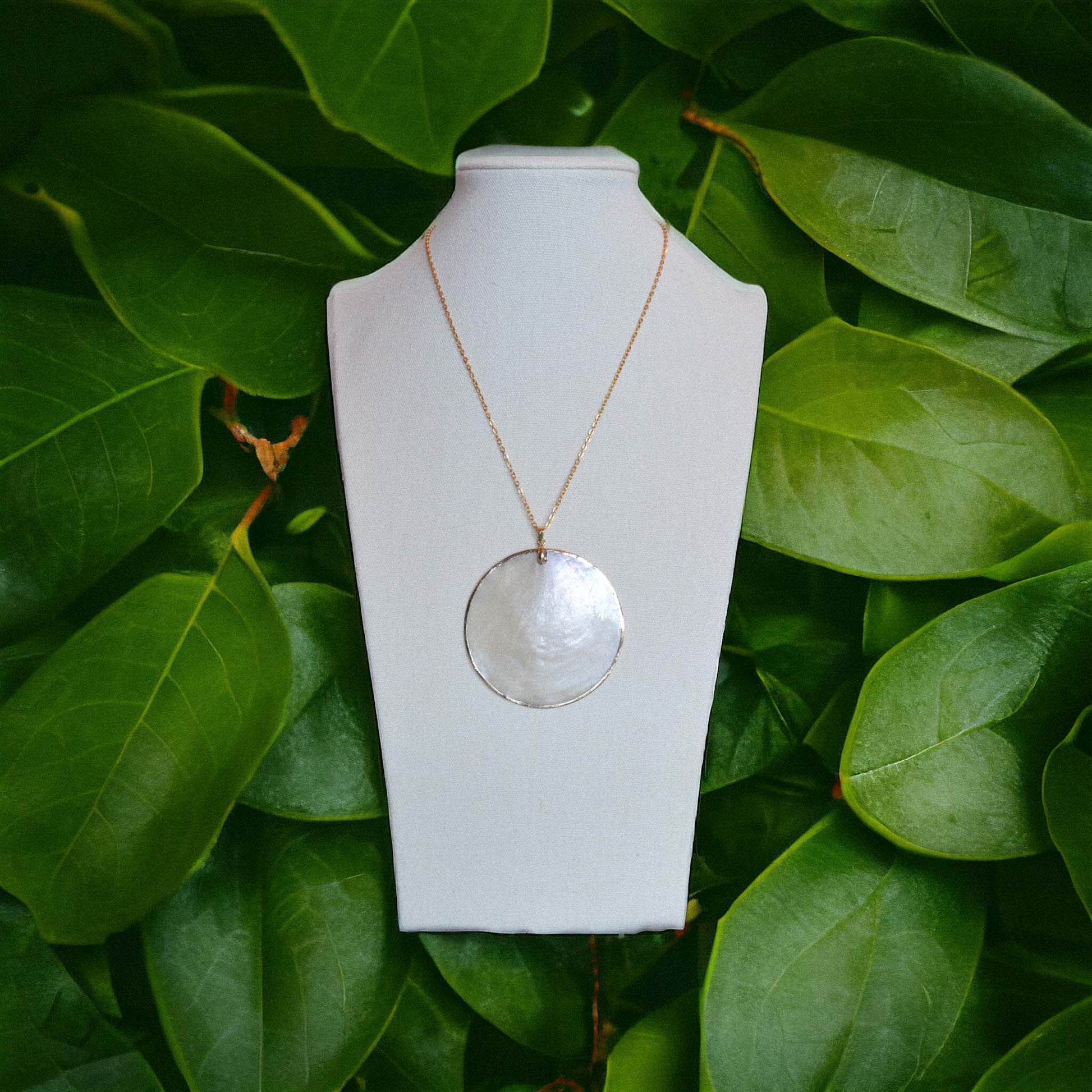 A statement gold-lined Capiz shell necklace displayed on a full-length wooden necklace holder, set against a lush backdrop of vibrant green tropical leaves.