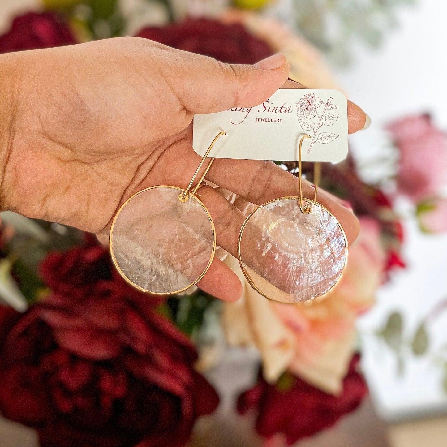A pair of Aking Sinta Zoey’s Capiz Shell Wedding Earrings; featuring large, oversized round white Capiz shell disks hanging from elegant gold hooks, displayed against a romantic, blurred red floral background.